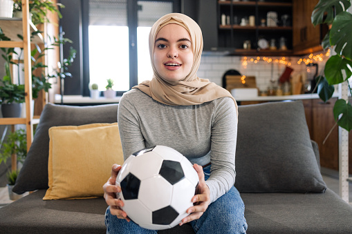 Portrait of a young Muslim soccer fan relaxing in the living room. She is sitting on the couch and holding a soccer ball while watching a soccer game attentively.