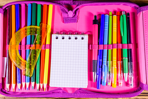 Different school stationeries (pens, pencils, notepad and protractor) in pink pencil box. Top view