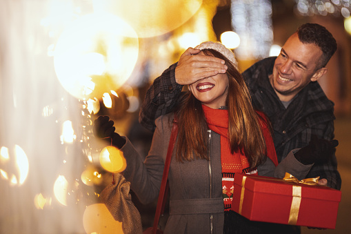 Young happy couple is walking along the city street with holiday bright in background. Smiling man is covering eyes of his happy girlfriend to surprise her with the gift in a Christmas night.