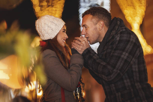 Young happy couple is walking along the city street with holiday bright in background. The boy is warming the smiling girl's hands with his breath.