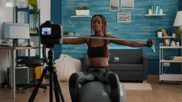 Athletic slim woman with black skin recording online yoga class using videocamera during fitness workout in living room. Fit athletic doing body exercices using aerobic elastic sitting on swiss ball