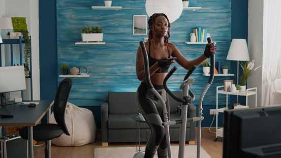 Cyclist woman with black skin in sportswear training body muscle using elliptical bicycle during bodyweight routine in living room warming up. Athletic slim fit adult enjoying healthy lifestyle