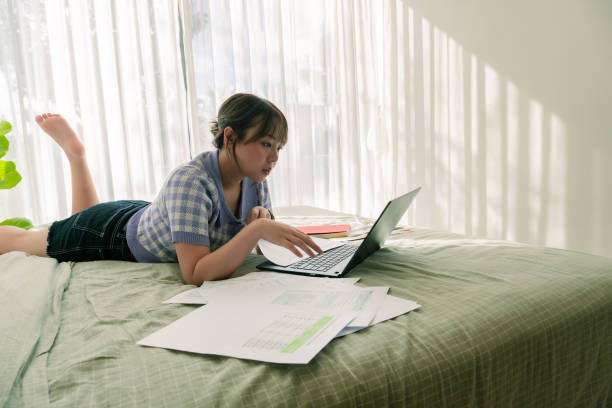 istock Asian financial advisor lying on front in a bedroom while looking at laptop for examining financial situation of a company, young adult female reviewing financial report and company budget in a laptop during sunny day 1729923455