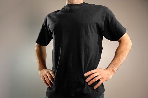 A close up crop of the torso of an anonymous caucasian man dressed in a plain black t shirt with his hands resting on his hips. The man is standing against a plain background. With copyspace for graphics to be added to the t shirt.