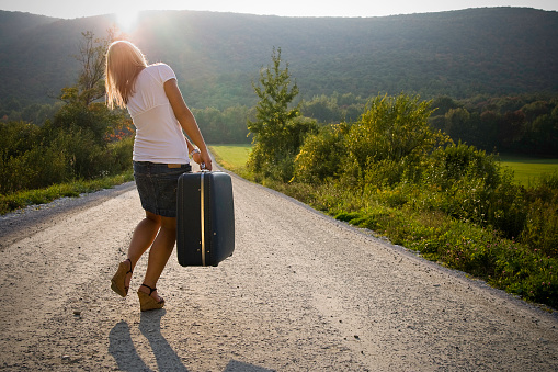 Young woman walking down rural road with heavy suitcase