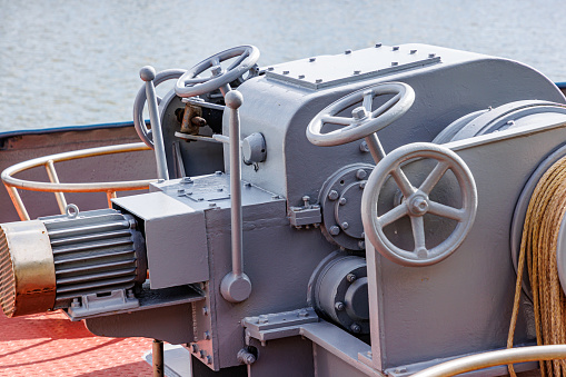 Closeup of levers, steering wheels, anchor windlass machine and heavy duty winches with tow rope at large mooring control station, bow of a cargo ship, blurred water in the background