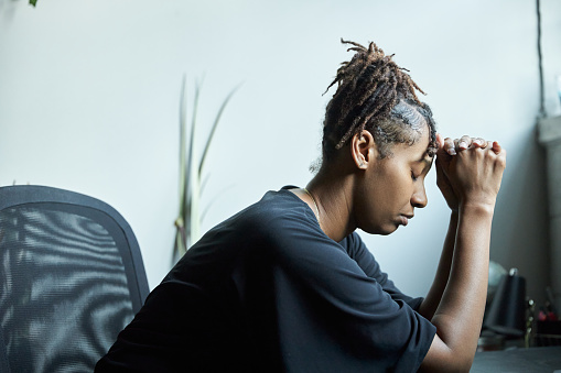 Young African-American woman clasping both hands to her head in distress while sitting at her desk at her home office with her eyes closed