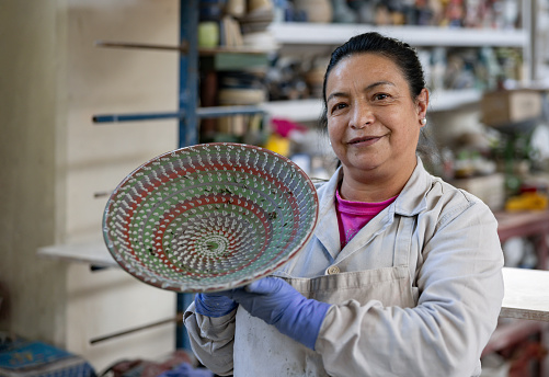 Portrait of a Latin American female potter working at a ceramics factory and looking at the camera smiling holding a bowl - manufacturing concepts