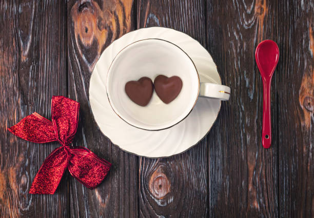 Concept for Valentine s Day. Chocolate hearts lie at the bottom of a white cup. Red bow and spoon as decor on a dark background.