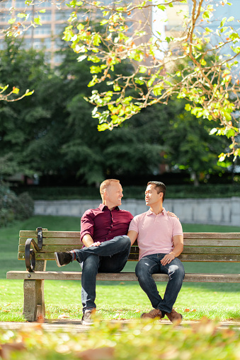 Middle aged gay couple, two men hugging, looking at each other, sitting in the park on bench. Love, date, relationship concept