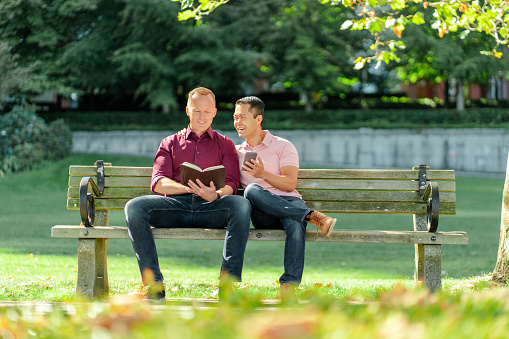 Portait of smiling homosexual men sitting on park bench reading book holding mobile phone outdoors. Concept of love, relationship, date
