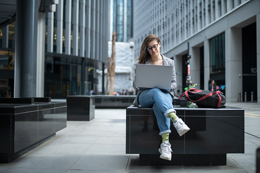 Confident young businesswoman working on laptop, sitting against corporate buildings in financial district in the city.