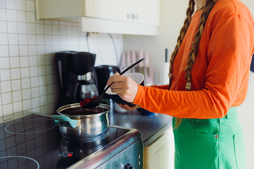 Female with long hair in colourful clothes cooking Swedish lingonberry jam in Scandinavian bright kitchen from berries harvested in the forest during autumn time