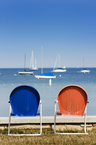 Red and blue lawn chairs overlooking sailboats on Lake Michigan beneath clear sky, Door County, Wisconsin, USA. Painted colors symbolic of liberal Democrats and conservative Republicans, the American pair sits side by side, ready for a political party debate, election discussion or tourist resort companionship. Copy space with no people.