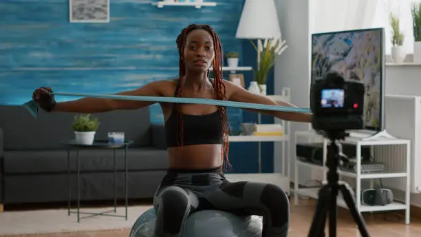 Trainer slim black woman recoring online yoga training using videocamera during morning fitness workout in living room. Fit athletic doing body exercices using aerobic elastic