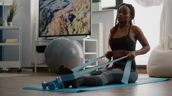 Athlete with black skin in sportswear exercising body muscle using fitness elastic enjoying healthy lifestyle sitting on yoga map in living room. Fit woman working at wellness warming before workout