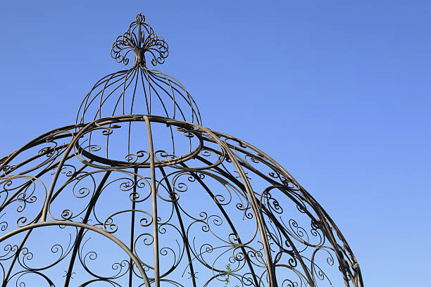 An ornate dome shaped garden gazebo, manufactured from bent metal. Isolated against a blue sky background.