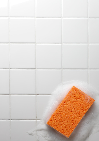 An orange sponge surrounded be soap suds sits on a grid of white bathroom tiles. There is ample room for copy. Image can be easily flipped horizontal to accommodate alternative composition needs.
