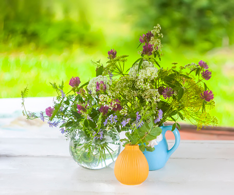 Photo of a china teapot being used as a vase, containing an arrangement of flowers. The teapot has a floral design and a gold trim and contains pink and white chrysanthemums and pink malva moschata. The vase stands on a table in a summery garden. Flowers can be seen in the background
