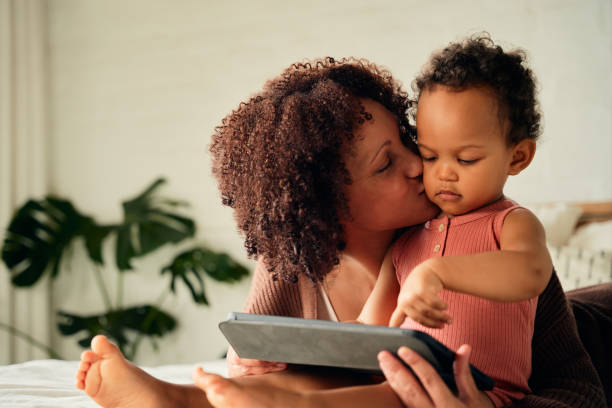 istock Biracial mother kissing daughter on cheek while using digital tablet on bed at home 1730020583