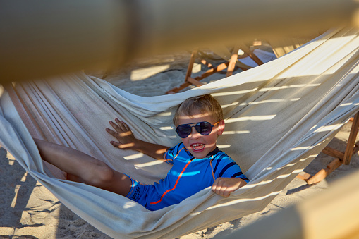 Carefree childhood of a cheerful boy on a summer vacation on the beach in a hammock. Beautiful cheerful little boy in sunglasses relaxes swinging in a hammock