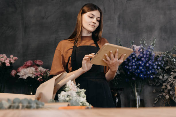istock Female florist accepts orders from customers for the creation of bouquets using digital tablet. Vertical photo. 1730070836