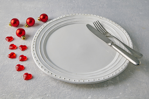 White ceramic plate with cutlery surrounded by red Christmas balls and hearts on a light concrete table. New Year's Food Display Template