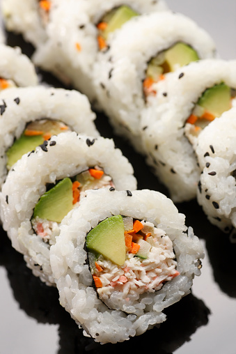 Close up of a row of California Roll sushi. A California Roll consists of crab, avocado, carrots, cucumber, wrapped in seaweed and sticky rice. Image is shot with a shallow depth of field with the focus centered on the closest roll.