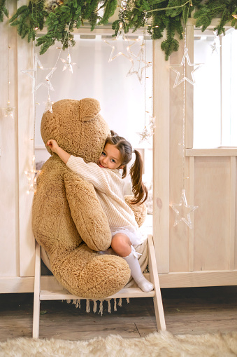A cute little girl hugs a big teddy bear sitting at a wall decorated with Christmas branches and garlands