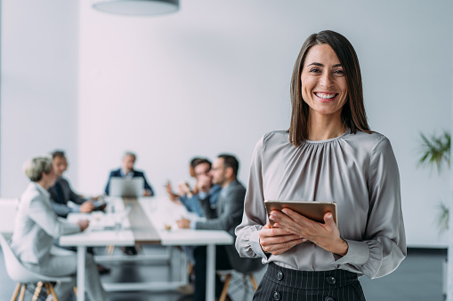 Shot of a beautiful smiling businesswoman standing in front of her team and holding digital tablet. Portrait of successful businesswoman standing with her colleagues working in background.