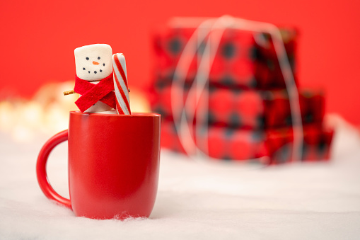 A marshmallow snowman in a red scarf smiling as he holds a peppermint stick in a mug of hot chocolate with a stack of Christmas presents in the background.