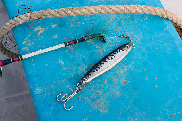 Close-up shot of the tip of a fishing pole laying on the blue seat of a boat with a shiny fish shaped jigging lure with a treble hook and a worn rope going across it. The boat seat has room for copy if you are looking for something to design.- A great shot for fresh seafood ingredient Photos or having to do with the fishing industry.