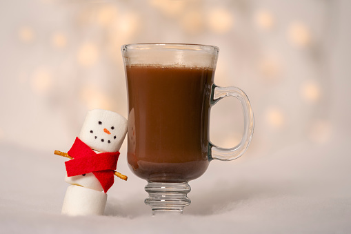 A marshmallow snowman in a red scarf smiling as he leans on a cup of hot chocolate.