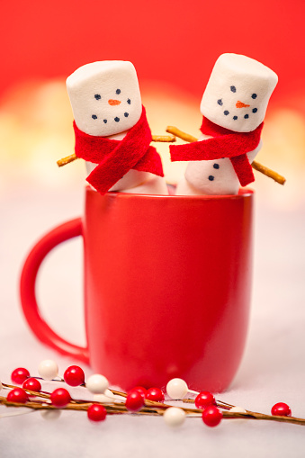 Two marshmallow men in red scarves smiling as they sit in a mug of hot chocolate photographed on a red background.