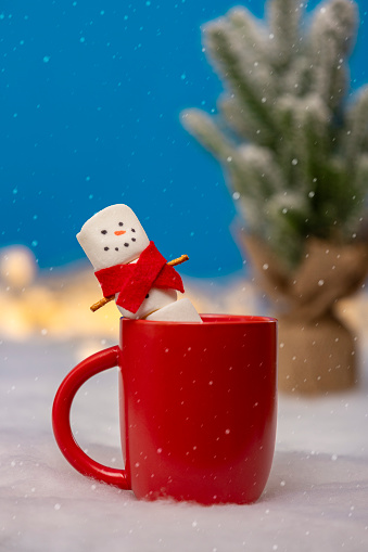 A marshmallow snowman in a red scarf smiling as he relaxes in a mug of hot chocolate.