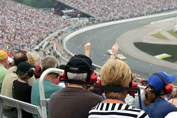 A DSLR photo of a couple of senior fans at a racing event. There is a crowd of people, the stadium is full and there is racing cars on the track. Some people are wearing headsets. There is also one person with arms raised in the air enjoying the race.