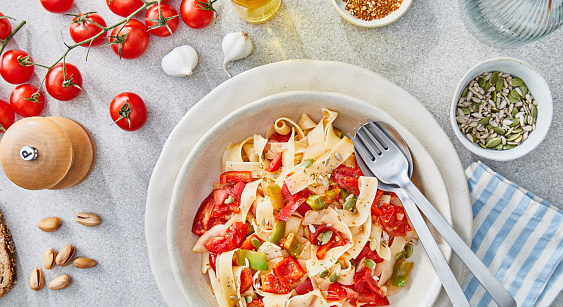 Delicious homemade autumn vegetarian pasta with fresh vegetables. red pepper, cherry tomato, green pepper, zucchini, served on a modern ceramic plate, with seasoning and herbs on a light rustic granite marble kitchen or restaurant table, representing a healthy lifestyle, wellbeing and body care, top view with a copy space