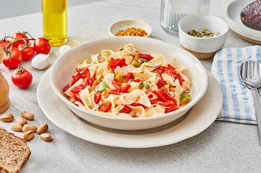 Delicious homemade autumn vegetarian pasta with fresh vegetables. red pepper, cherry tomato, green pepper, zucchini, served on a modern ceramic plate, with seasoning and herbs on a light rustic granite marble kitchen or restaurant table, representing a healthy lifestyle, wellbeing and body care, top view with a copy space