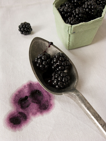 Some fresh blackberries in a spoon with a blackberry stain on the tablecloth.