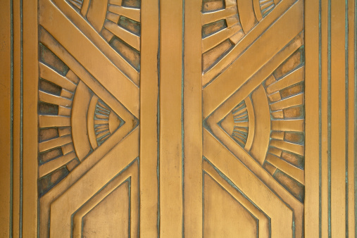   Close-up architectural detail from an art deco style bronze door.  The photo was taken in New York City and the composition focuses on the strong vertical elements juxaposed with radiating diagonals suggesting the rays from the sun. The detail is rich with golden tone hues and patination conducive to a building from 1929.