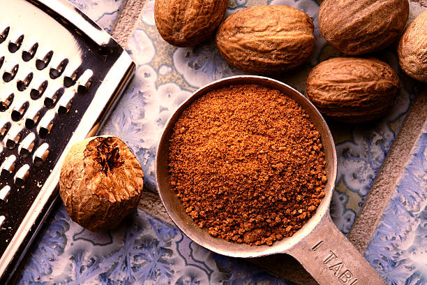 Ground nutmeg in measuring spoon Ground nutmeg in measuring spoon, with un-ground nuts and grater on patterned blue tile ground. nutmeg stock pictures, royalty-free photos & images