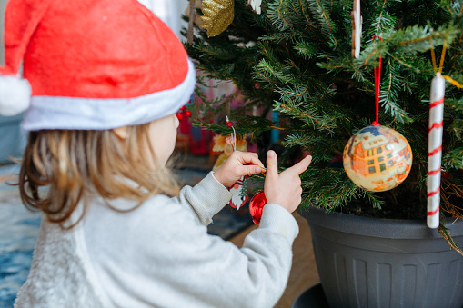 rear view on little girl with light hair in santa hat holding red christmas tree decoration in hands putting it on tree