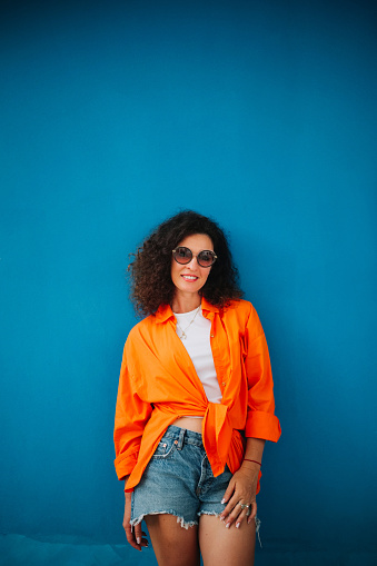 Bright and colorful. Stunning curly haired woman in shorts and orange shirt posing against violet wall.