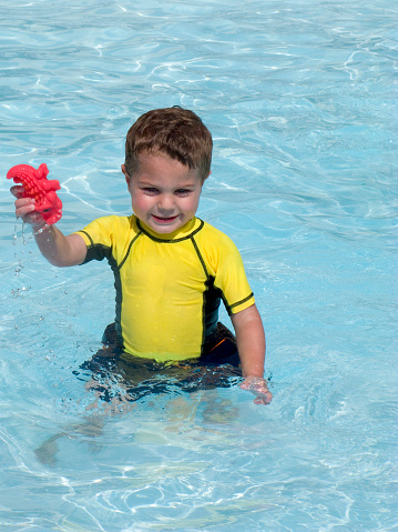 Shown here is a two-year-old toddler playing in the shallow end of a swimming pool with some generic plastic pool toys.  He is wearing a bright yellow sun shit for protection from the sun.