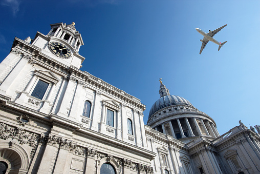 An airliner flies at low altitude over London's iconic St Paul's Cathedral on a bright sunny day.