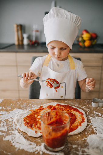 Adorable smiling 3 years old girl in chefs hat and apron cooking pizza at the kitchen, puts tomato sauce. Kids hobbies