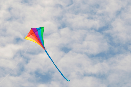 A rainbow colored delta kite sails on a cotton candy cloudy sky