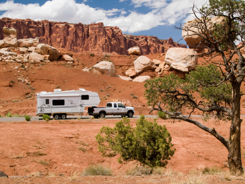 A Fifth Wheel Travel Trailer ( 5th Wheel Trailer ) on a paved road with a rocky brown colored hillside and a green tree framing the foreground. Located at Capitol Reef National Park, a United States National Park, located in south-central Utah.