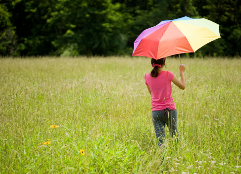 Rear view of teen girl walking in field of grass holding multicolored umbrella