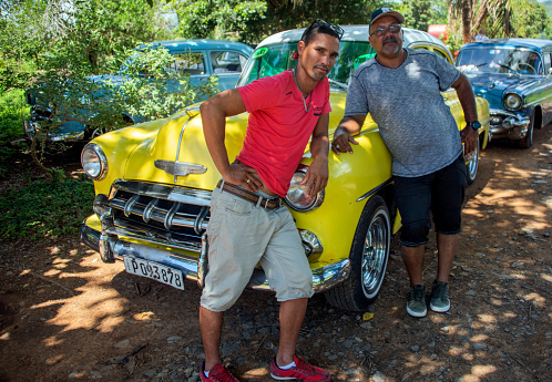 Pinar del Rio, Cuba - 6/28/17: Two taxi drivers lean on a 1950s-style shiny yellow Chevrolet. Other American-made cars are parked in the dirt lot in the Cuban province of Pinar del Rio.